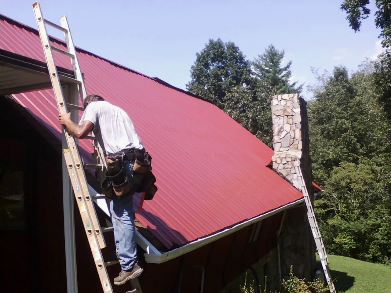 Roofer on a ladder beside a red metal roof and stone chimney — Soffit & Fascia Repair by Roofing Brothers in Emlenton